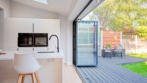 Een moderne keuken met witte kasten en een zwarte afzuigkap, een witte stoel en een schuifdeur naar een buiten terras met een houten tafel en stoelen.