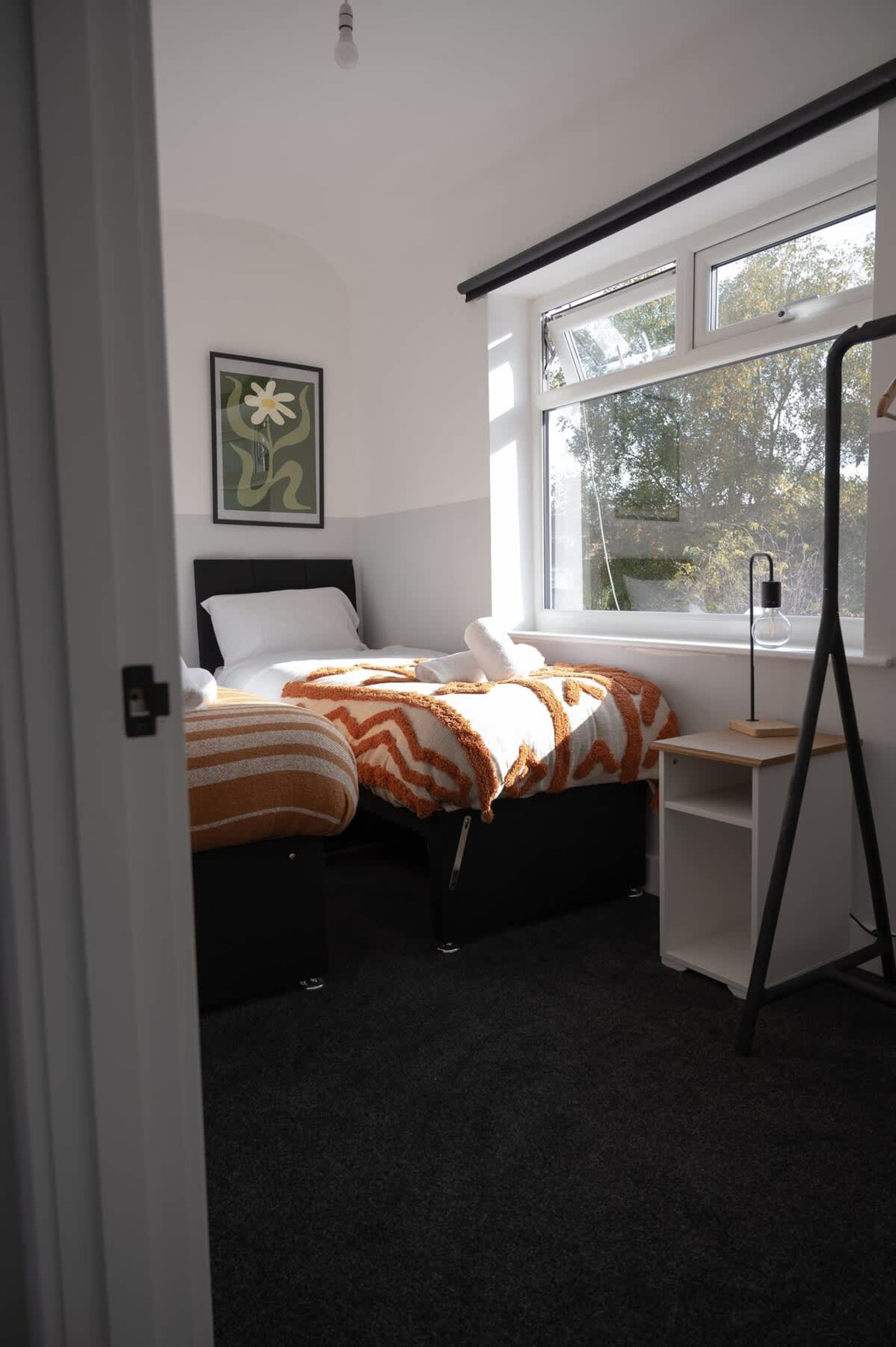 Un dormitorio con dos camas, un escritorio, una lámpara de pie y una ventana con vistas a árboles.