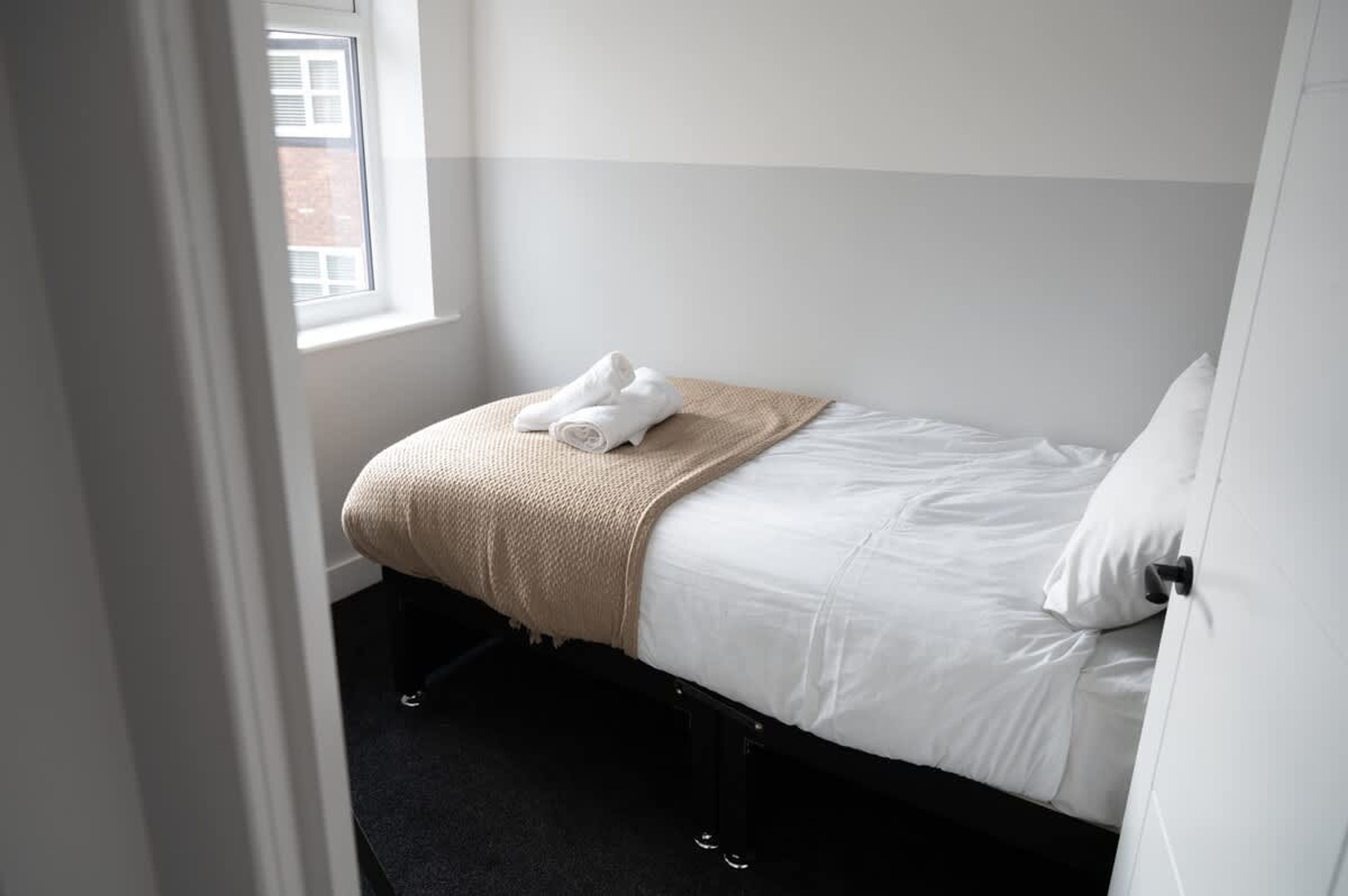 Ein Einzelbett mit weißer Bettwäsche und einer beigen Tagesdecke, ein Fenster mit Jalousien und eine Tür links.