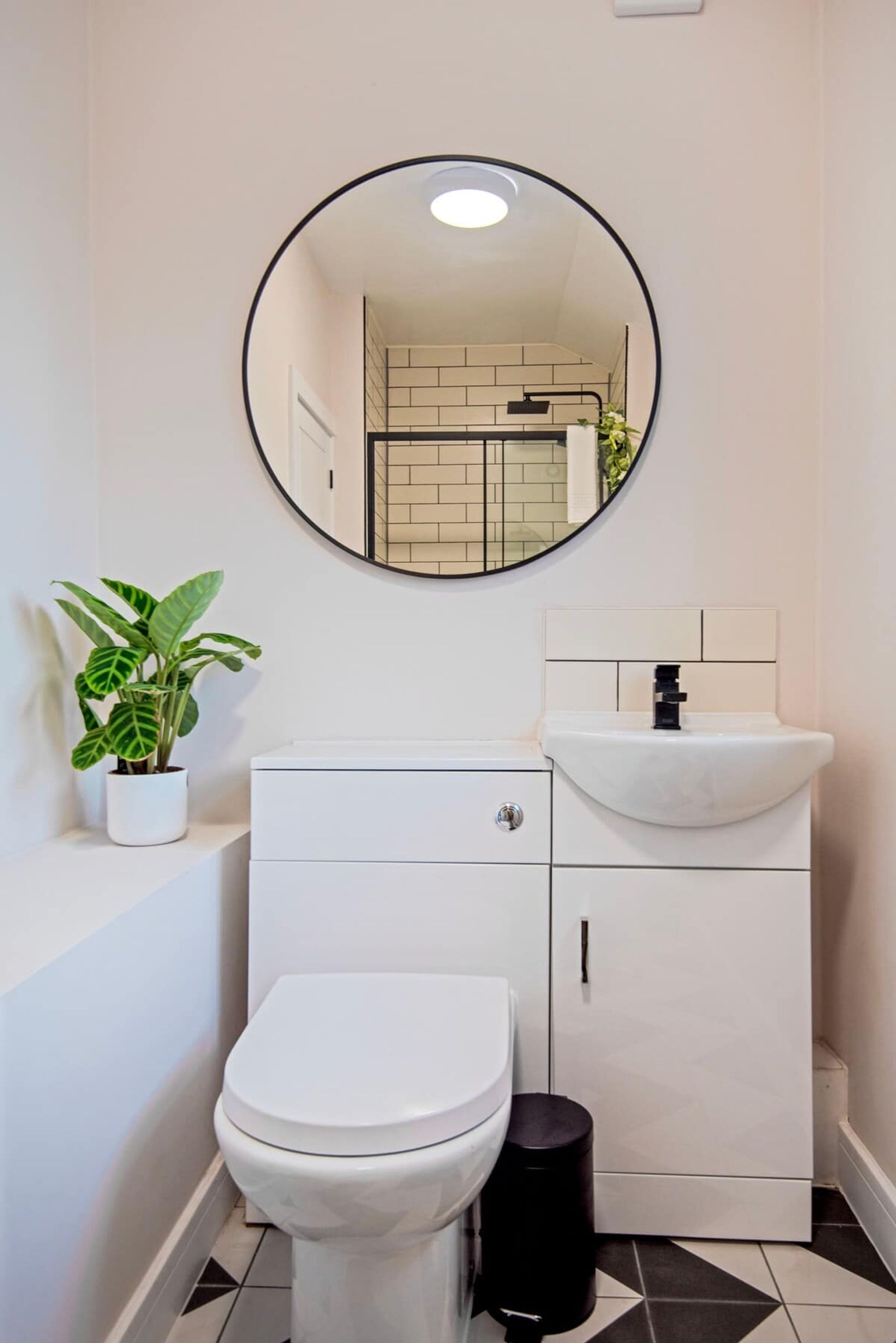 Ein modernes Badezimmer mit Toilette und Waschbecken in Weiß, einem runden Spiegel und einem Blumentopf.