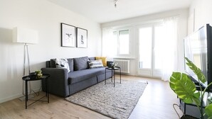 A modern living room with a grey sofa, a small side table, a floor lamp, and a large window with white curtains. - Modern Flat w Netflix Balcony top Location (Lucerne)