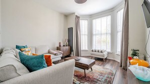 A modern living room with a sofa, coffee table, and a TV. There are windows with curtains, a radiator, and a plant. - Cosy Apartment in Stunning Didsbury (Manchester)