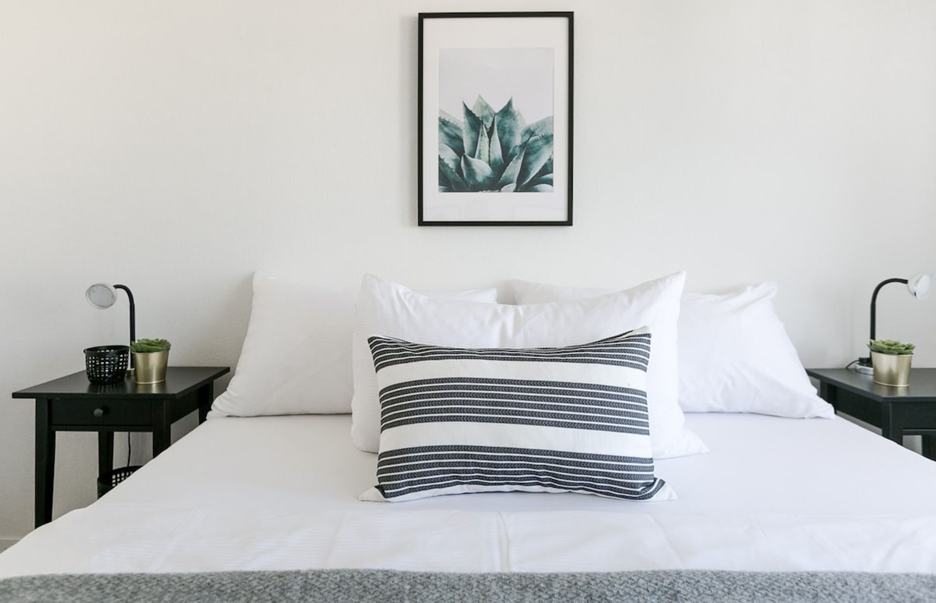 A neatly made bed with white bedding and striped pillows, a framed plant on the wall, and two bedside tables with lamps and small potted plants.