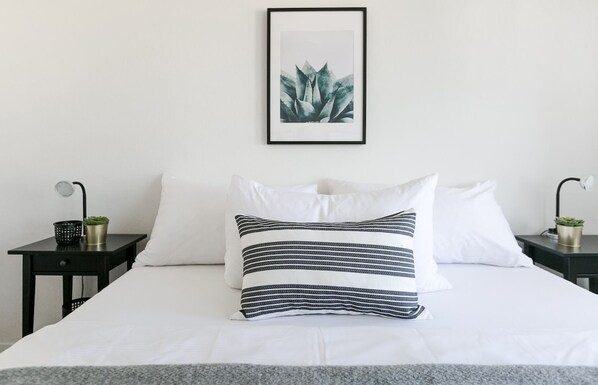 Una cama bien hecha con ropa de cama blanca y almohadas a rayas, una planta en un marco en la pared y dos mesitas de noche con lámparas y pequeñas macetas.
