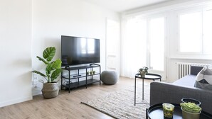 A modern living room with a flat-screen TV on a black stand, a grey sofa, a round ottoman, and a small side table with a plant. - Lucern 2-br Flat w Netflix Top Location (Lucerne)