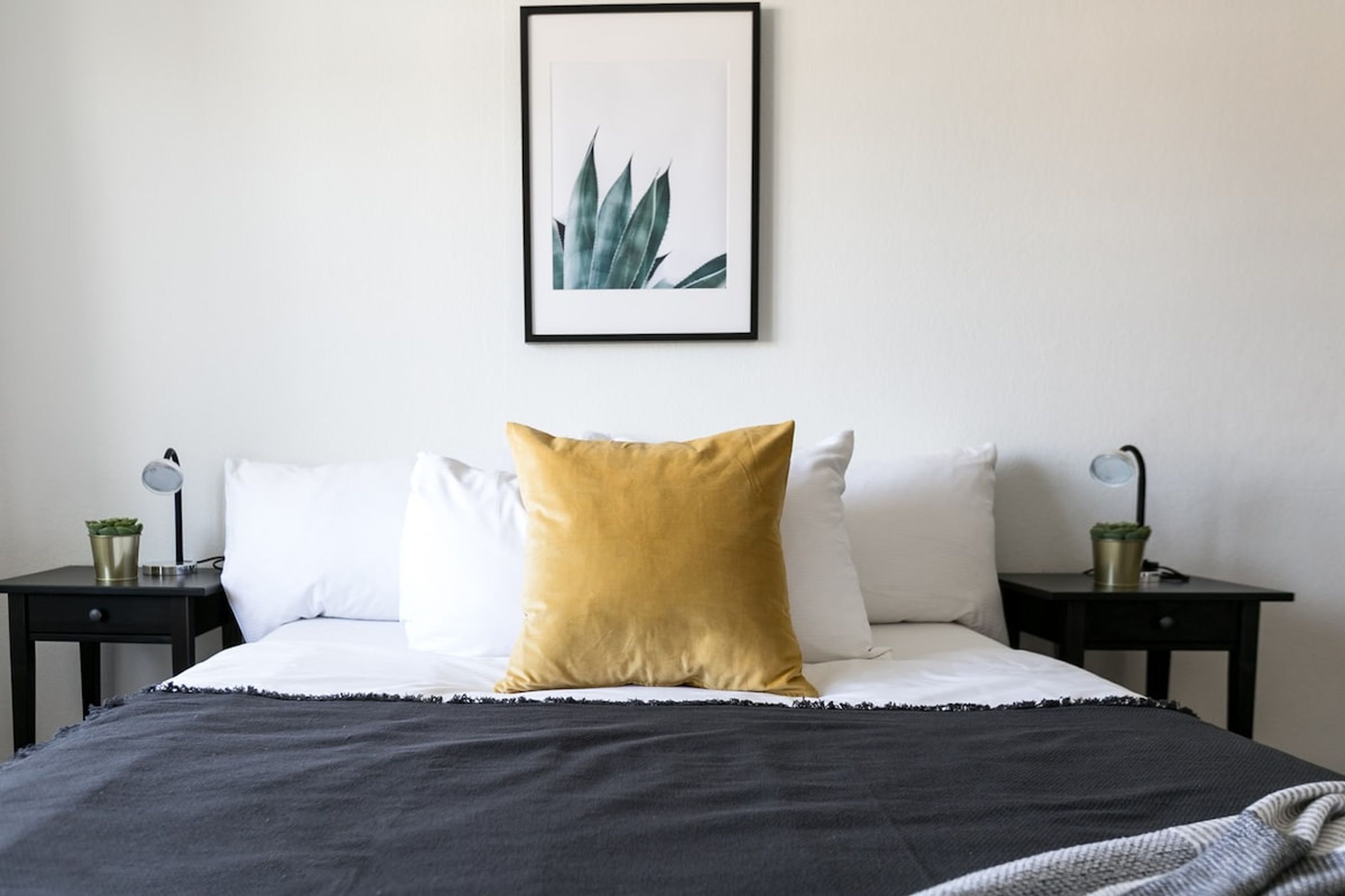 Una cama bien hecha con un edredón blanco y negro, una almohada amarillo mostaza y una planta enmarcada en la pared.