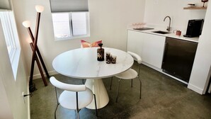 Una cocina compacta con encimera blanca, un lavamanos y una cafetera. Hay una mesa de comedor con cuatro sillas y un florero decorativo.