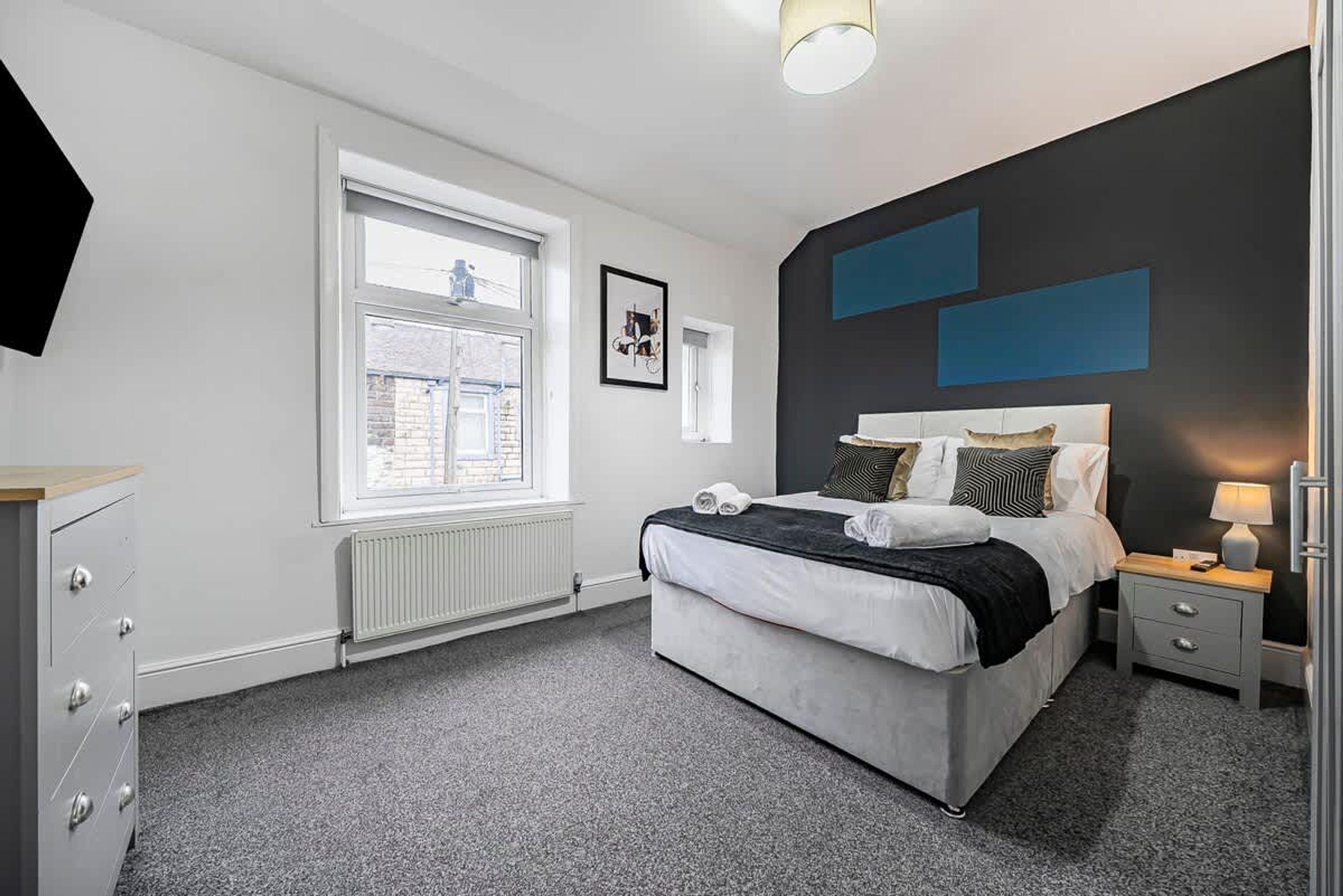 A modern bedroom with a large bed, a dresser, a window, and a wall with blue panels.