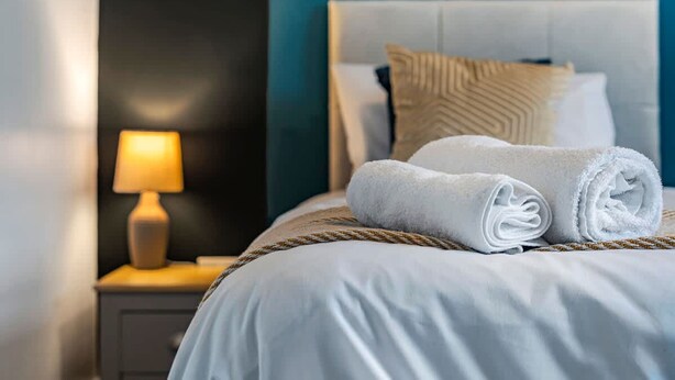 A neatly made bed with white linens and rolled towels, a bedside lamp, and a headboard with decorative pillows.