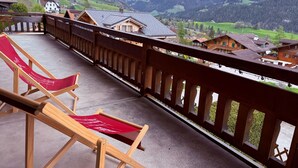 Un balcón con barandillas de madera, dos sillones reclinables rojos y vistas a un paisaje montañoso con casas.