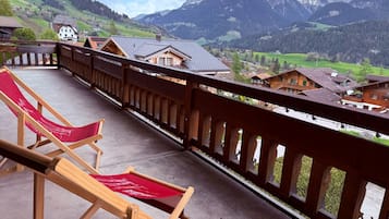 Ein Balkon mit Holzbalkonen, zwei rote Liegestühle und Blick auf eine Berglandschaft mit Häusern.