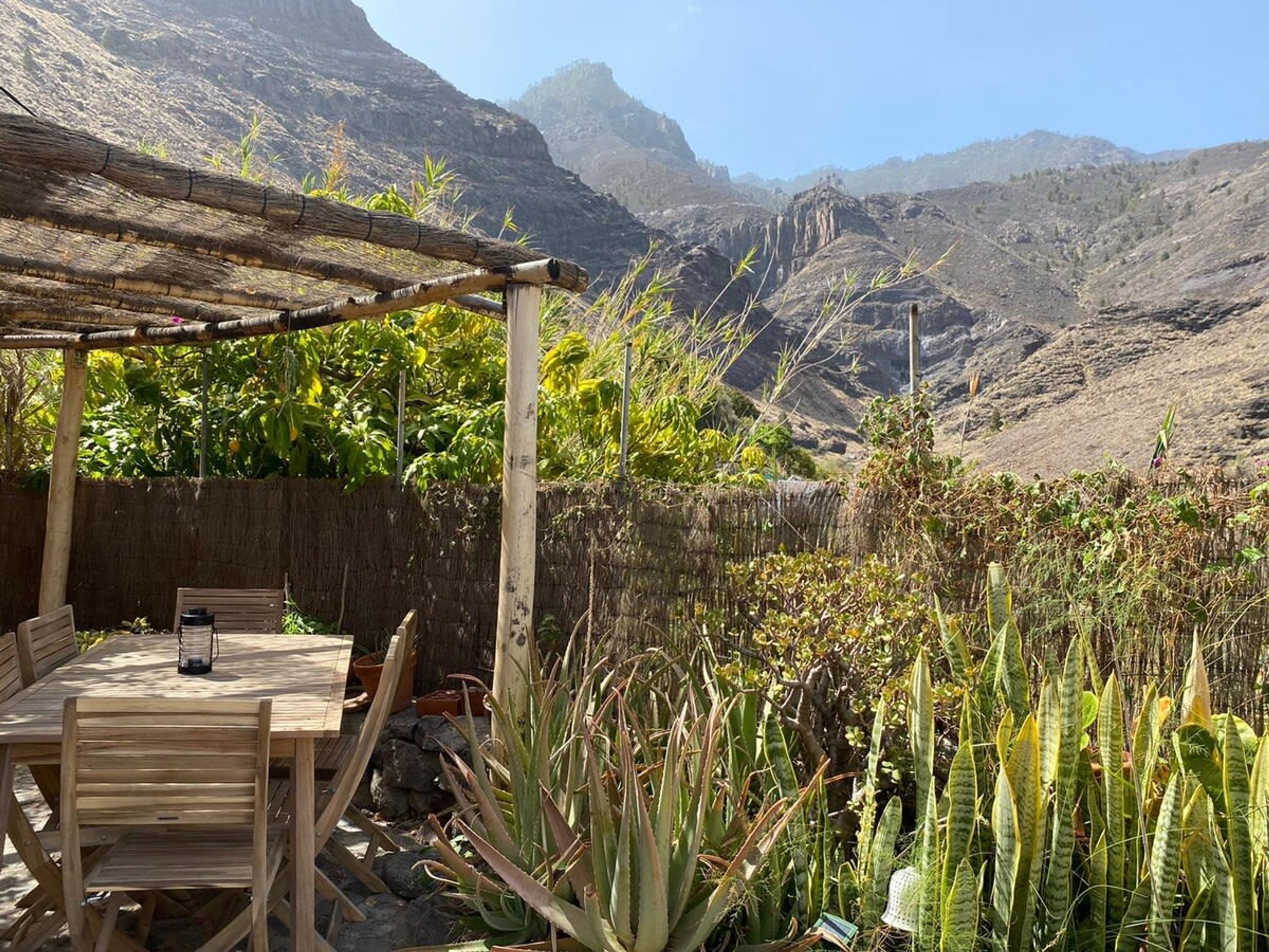 Eine Holzpergola mit Tisch und Stühlen, umgeben von Kakteen und Pflanzen, mit Blick auf eine Berglandschaft.