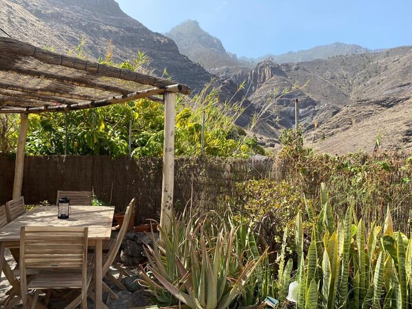 A wooden pergola with a table and chairs, surrounded by cacti and plants, overlooking a mountainous landscape.