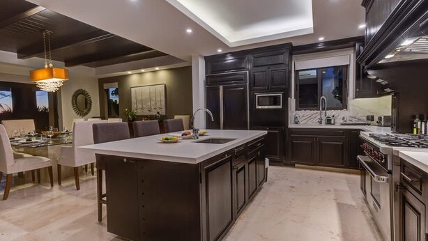 Una cucina moderna con mobili scuri, una grande isola e un'area da pranzo con un tavolo e sedie in vetro.