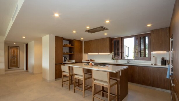A modern kitchen with wooden cabinets, a central island with bar stools, recessed ceiling lights, and a large window with blinds. - Encanto 822 by Owner Luxury Sunset Getaway Punta Mita (Punta de Mita)