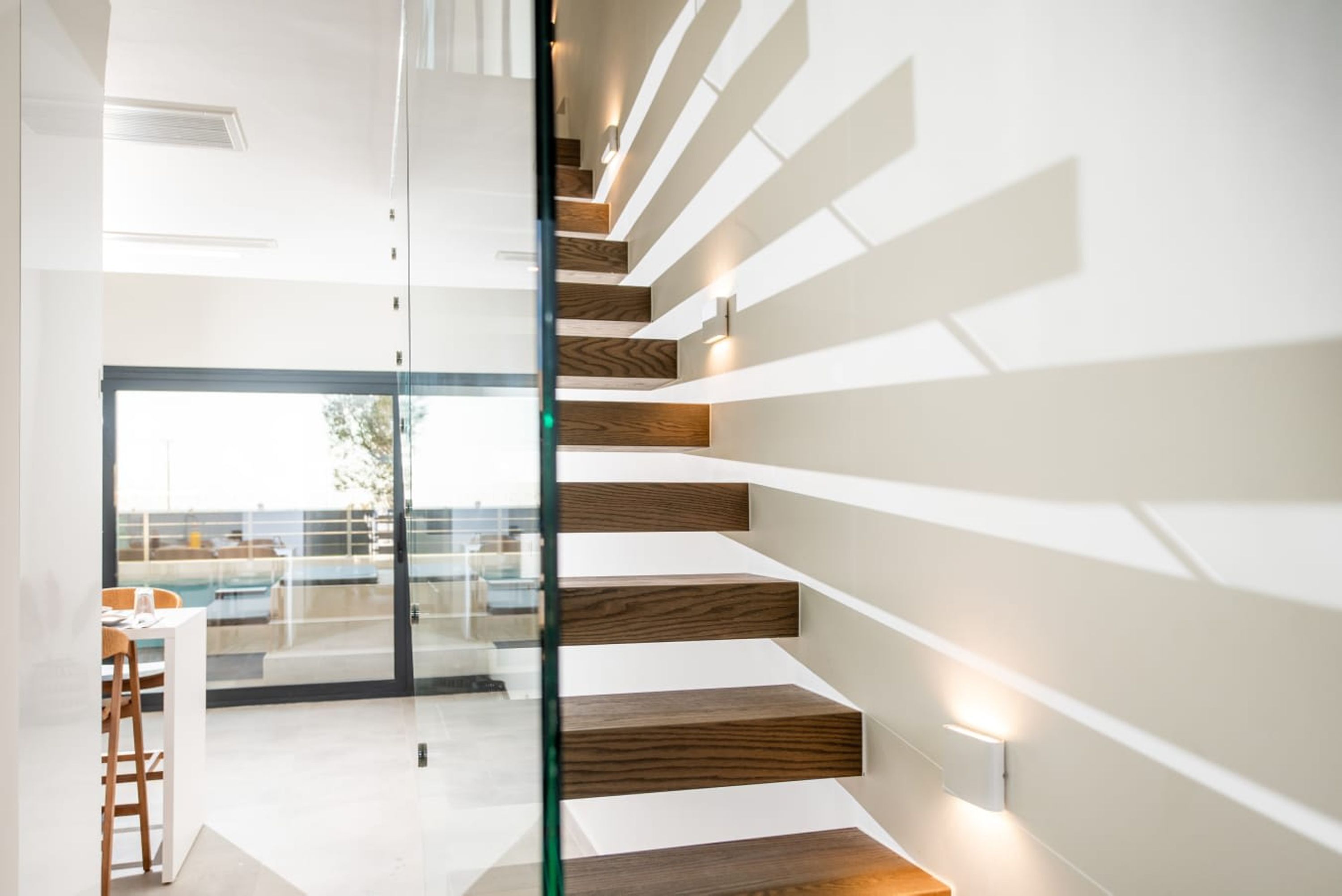 A modern staircase with wooden steps and glass railings, leading to a balcony with a view of the outdoors.