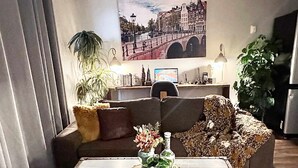 Un saló amb una taula de centre de velles baules, un sofà amb coixins, un escriptori amb un ordinador i un gran quadre de paisatge urbà a la paret.