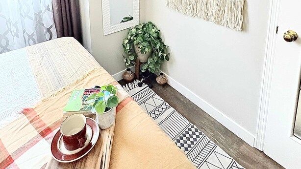 Una habitació amb un llit, una taula petita amb una tassa i un plat, una planta i un penjador de macramé a la paret.