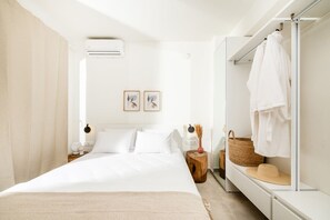 A minimalist bedroom with a large bed, a bedside table with a lamp, a mirror, and a wardrobe with hanging clothes.