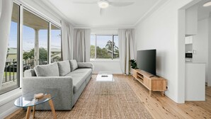 Una sala de estar moderna con un sofá gris, una mesa de centro de cristal, un televisor de pantalla plana y un amplio ventanal con cortinas.
