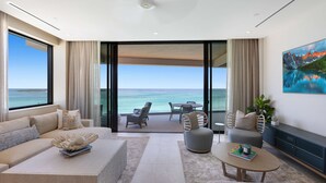 A modern living room with a sofa, coffee table, and chairs, a balcony with a view of the ocean, and a painting on the wall.