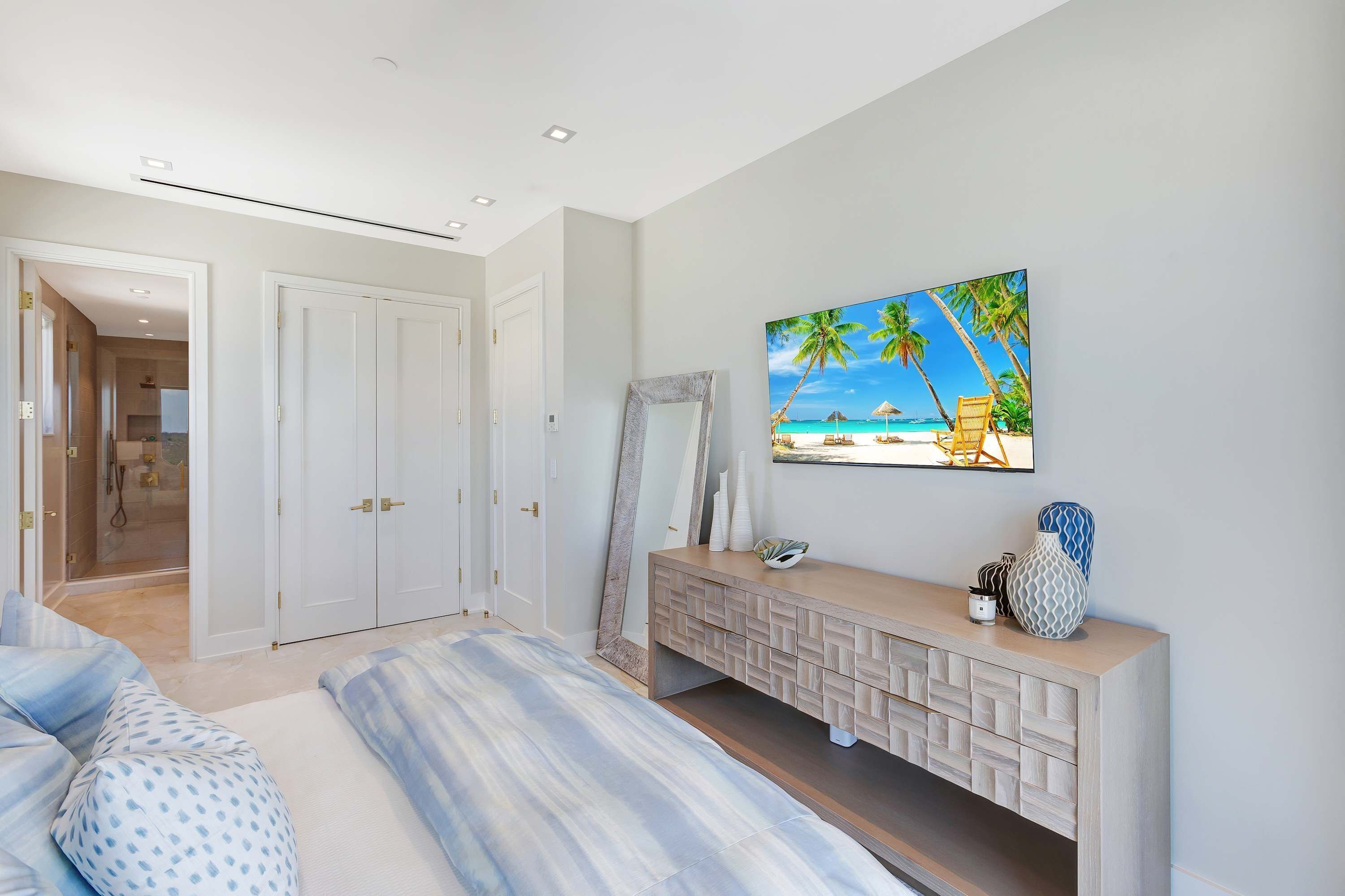 A bedroom with a bed, a vanity with decorative items, a painting of a beach scene, and a shower area.