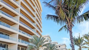 A modern hotel with a curved facade, multiple balconies, and a well-maintained garden with palm trees and a pathway.