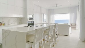 A modern kitchen with white cabinetry, a central island with bar stools, and a view of the sea through large windows. - Lilypad Luxurious Cliffside Villa With Stunning Sea Views (Ocean City)