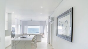 A modern kitchen with a marble countertop, a dining area with white furniture, and a large window overlooking the sea. - Lilypad Luxurious Cliffside Villa With Stunning Sea Views (Ocean City)