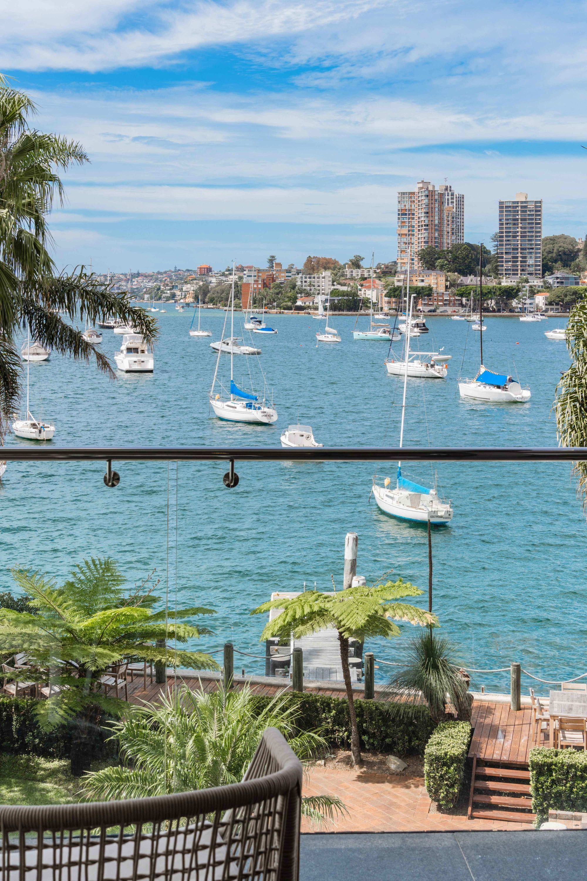 A marina with multiple sailboats docked, a wooden deck with outdoor seating, and buildings in the background.