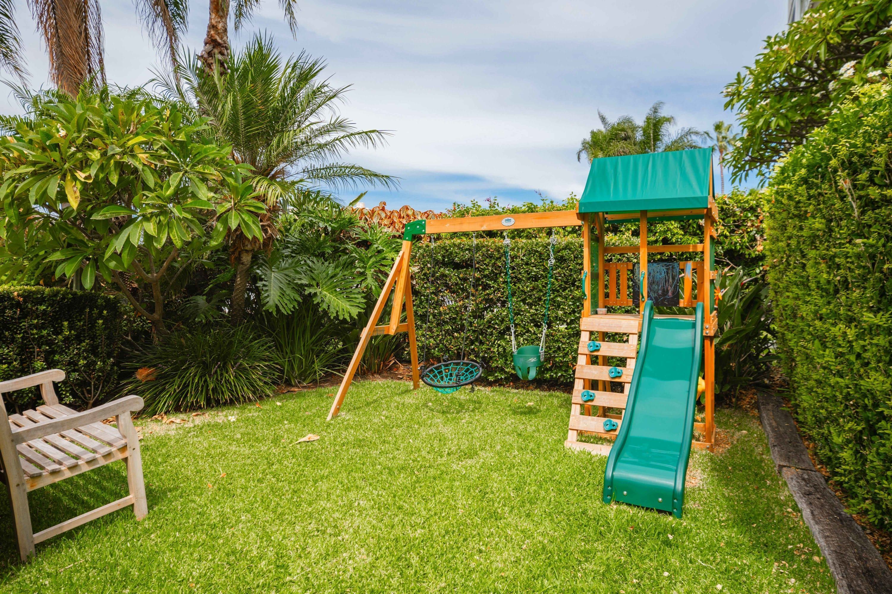 A wooden swing set with a green slide, surrounded by lush greenery and a bench.
