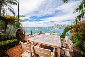 A wooden deck with a table and chairs overlooking a marina with boats and buildings in the distance.