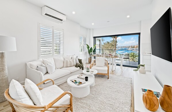 A modern living room with a white sofa, wicker chairs, a coffee table, and a large window offering a marina view.