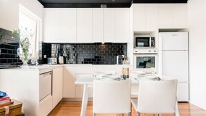 A modern kitchen with white cabinetry, a black tiled backsplash, a dining table with white chairs, and a white refrigerator. - Renovated Retro Apartment Near Harbour Front Walks (Sydney)