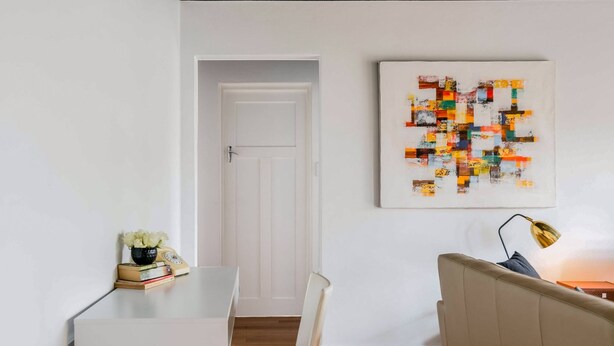 A modern living room with a white sofa, a small table with books and a vase, and a colorful abstract painting on the wall. - Renovated Retro Apartment Near Harbour Front Walks (Sydney)
