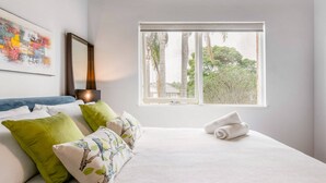 A neatly made bed with white bedding and green and floral pillows, a large window with a view of trees and buildings, and a framed abstract painting on the wall. - Renovated Retro Apartment Near Harbour Front Walks (Sydney)