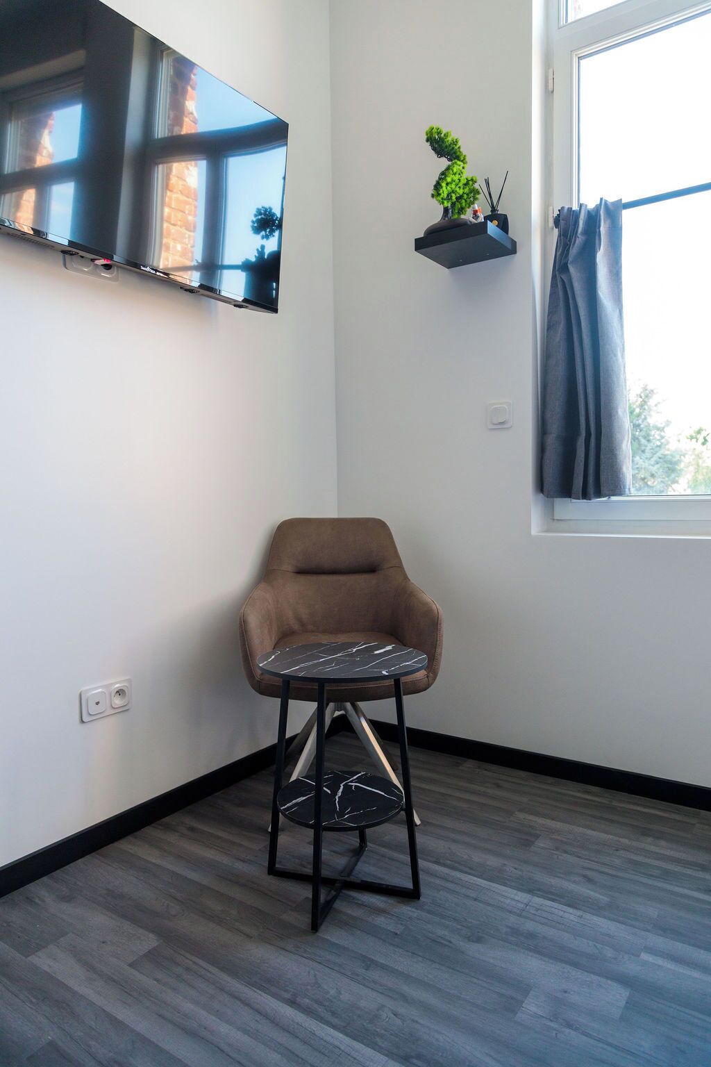 Een moderne woonkamer met een bruine leren fauteuil, een klein zwart-wit marmeren tafeltje, een wandgemonteerde flatscreen televisie en een raam met gordijn.