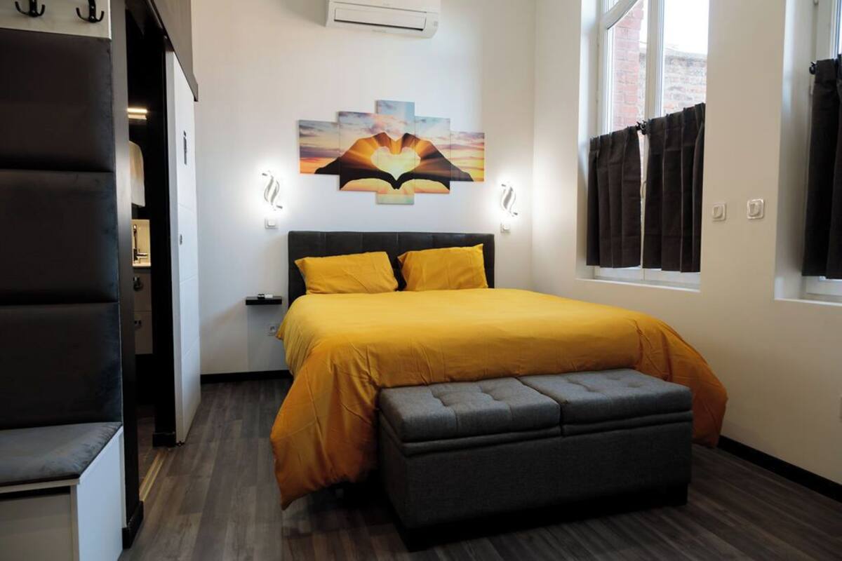 Een slaapkamer met een bed, een bankje, een kunstwerk aan de muur en een raam met gordijnen.