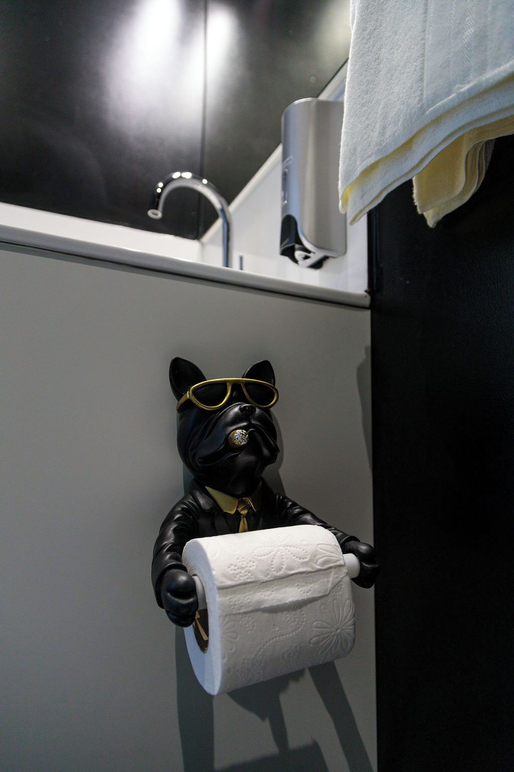 Un baño con un dispensador de papel higiénico con forma de perro de gafas de sol, una toalla colgada en la pared y un dispensador de papel.