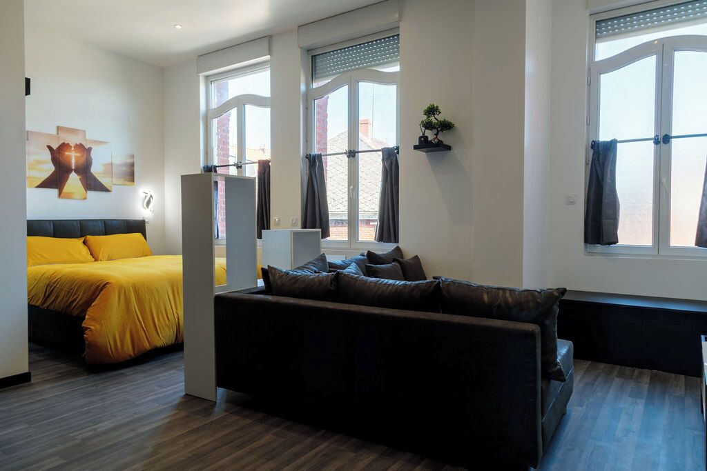 A modern living room with a sofa, a bed with yellow bedding, and large windows with curtains.