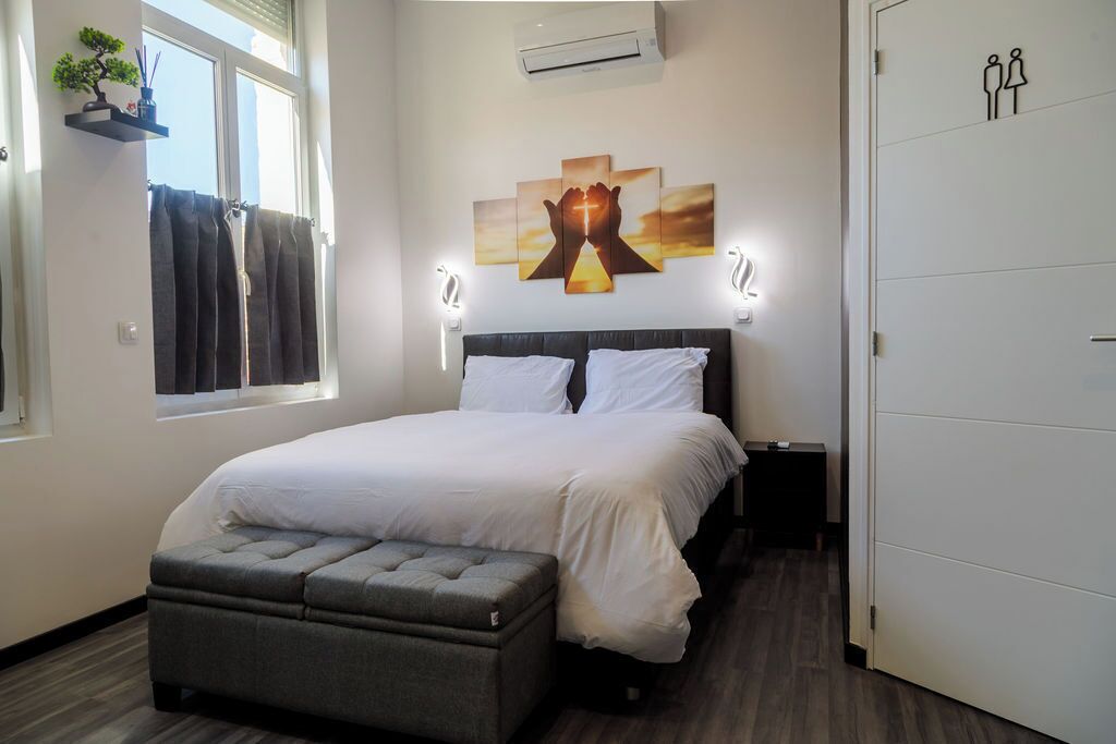 A bedroom with a bed, bedside tables, a window with curtains, a wall-mounted air conditioner, and a door with a gender symbol.