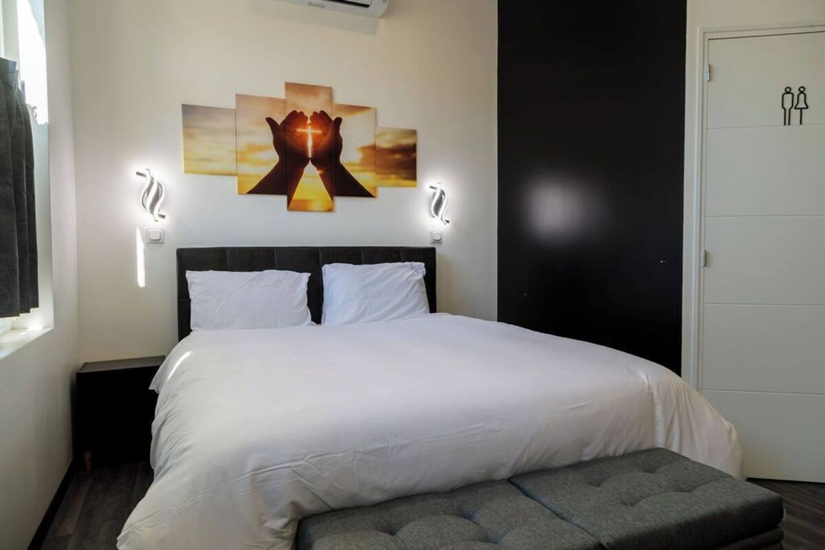 Een net opgemaakt bed met witte lakens, een grijs gestoffeerde bank en een wanddecoratie van handen die een kruis vasthouden.