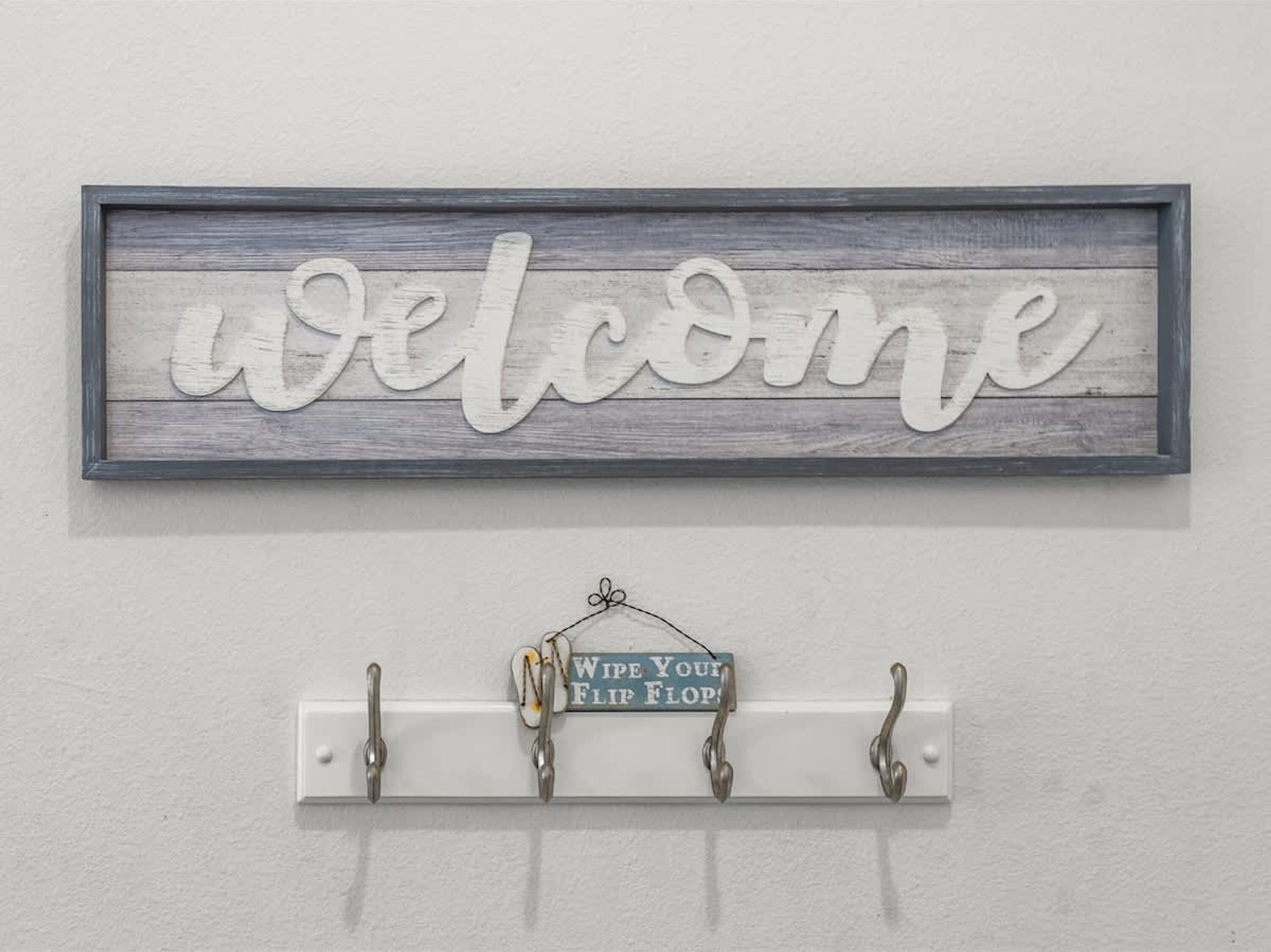 A 'welcome' sign with a rustic wooden background and a metal sign below with hooks and a 'wipe your flip flop' sign.