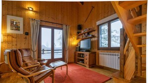 Interior de una cabaña de madera con una butaca de cuero, televisor, chimenea y vistas a montañas nevadas.