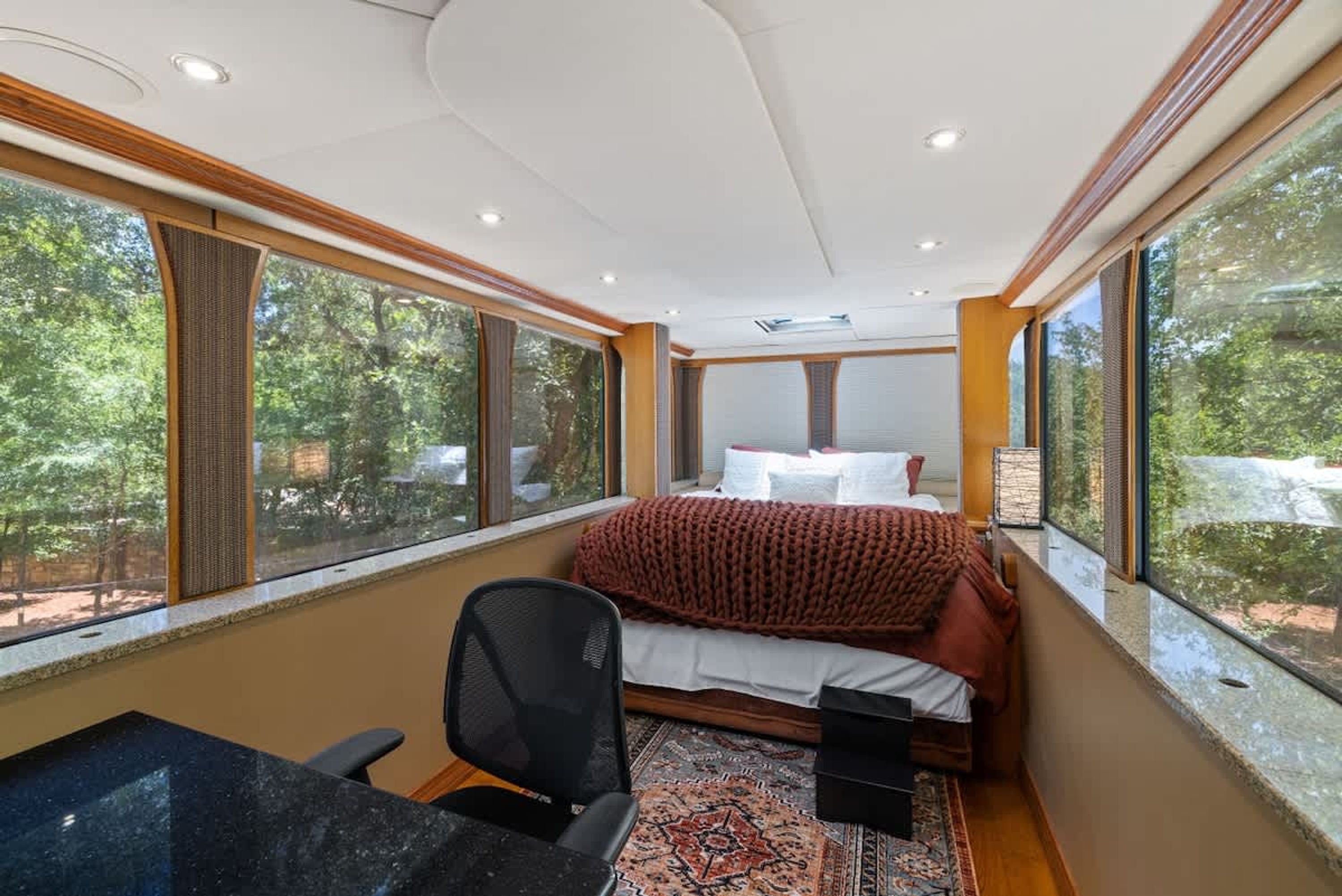 Une chambre à coucher avec un lit, un bureau et une chaise. Il y a de grandes fenêtres d’un côté.