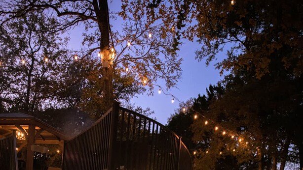 Un chemin de pierres menant à une terrasse en bois illuminée de guirlandes lumineuses, entourée d’arbres et d’un mur de pierres.