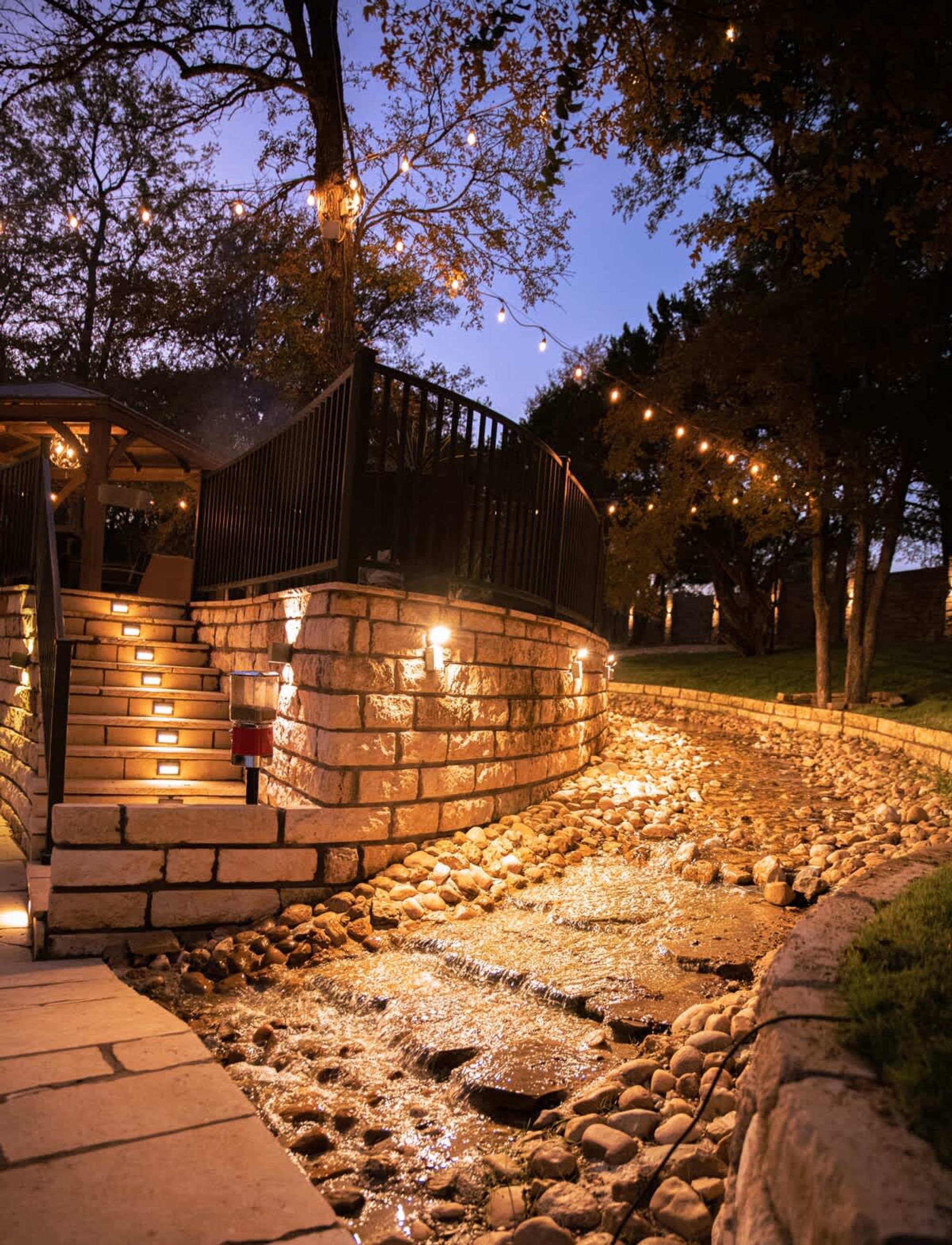 A stone pathway leading to a wooden deck with string lights, surrounded by trees and a stone wall.