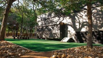 A building with a green artificial turf lawn, surrounded by trees and a gravel path.