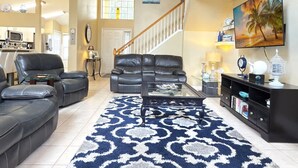 A spacious living room with a large black leather sofa, a coffee table, and a patterned rug. A staircase with a wooden railing leads to an upper level. - Near Disney Villa 3 bd Private Pool (Davenport)