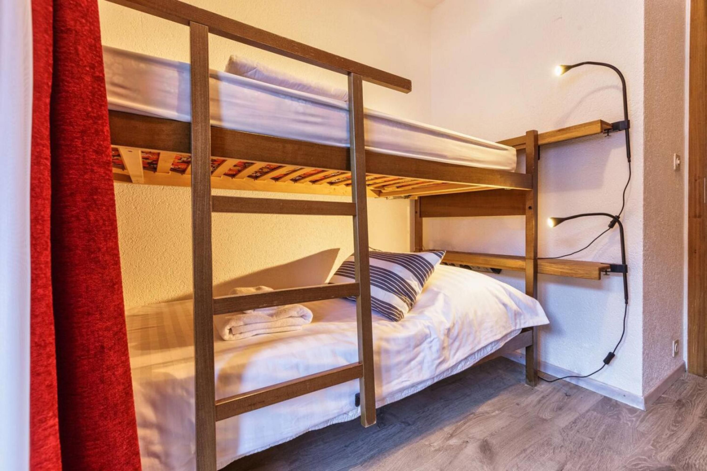 A bunk bed with a wooden frame, a white mattress, and a striped pillow. There is a red curtain partially visible on the left.